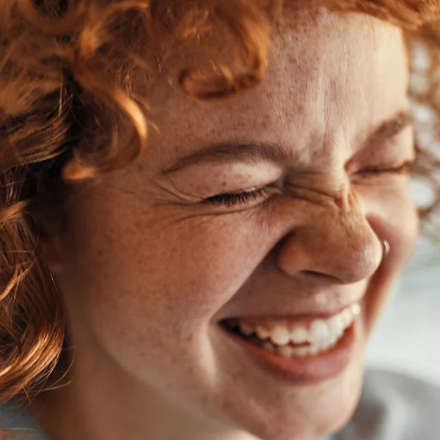 Close-up of a joyful woman with red, curly hair and freckles laughing heartily